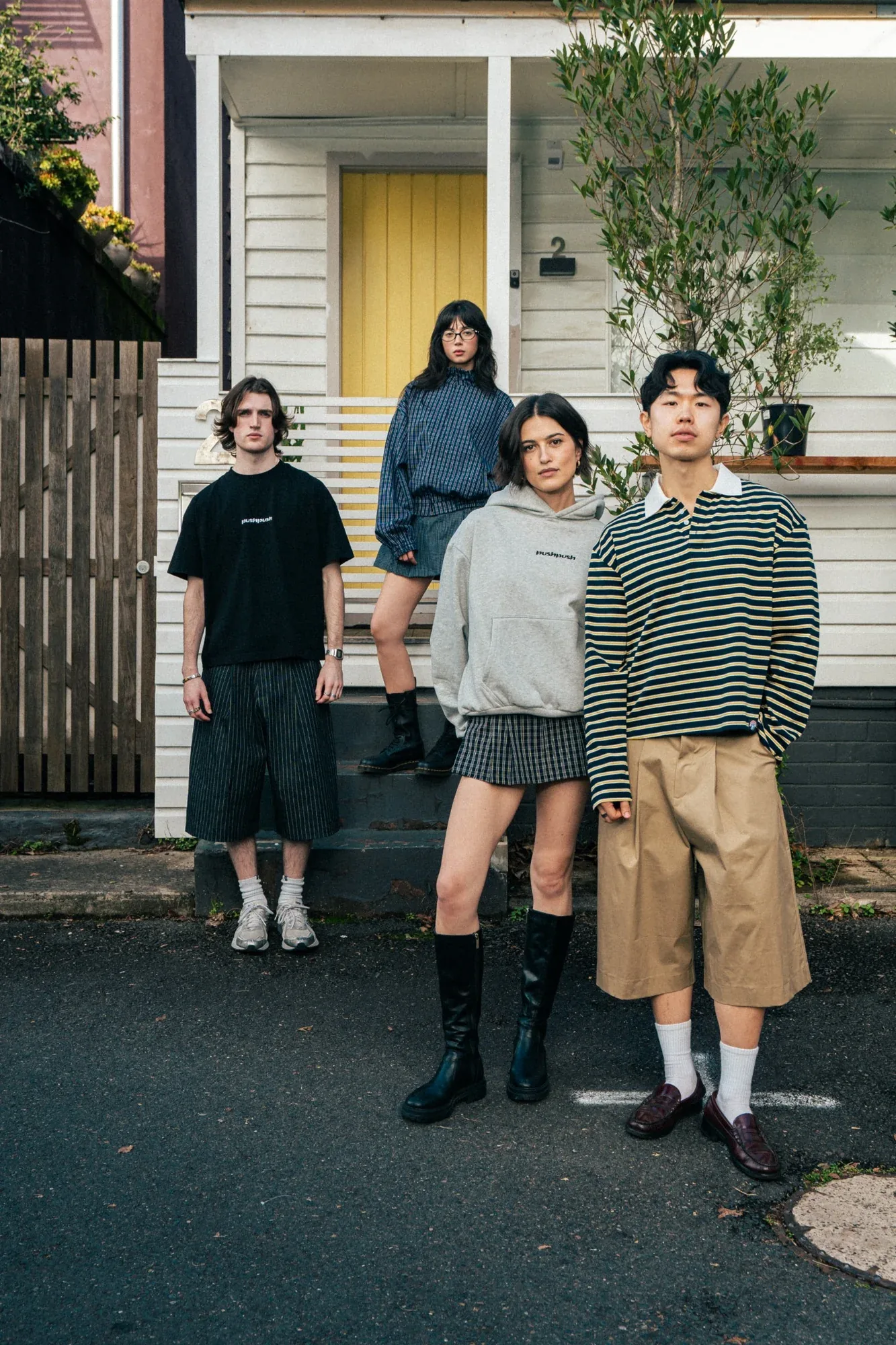 Five models on a Sydney terrace street, a yellow front door behind: oversized tees, a grey hoodie, a striped knit, pleated skirts and wide shorts