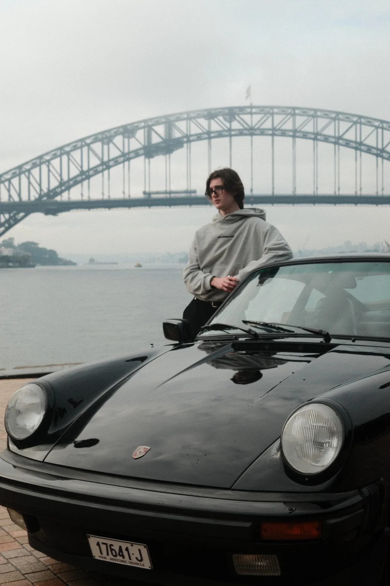 A model in a grey hoodie leans on a black Porsche 911 by the water, the Sydney Harbour Bridge behind
