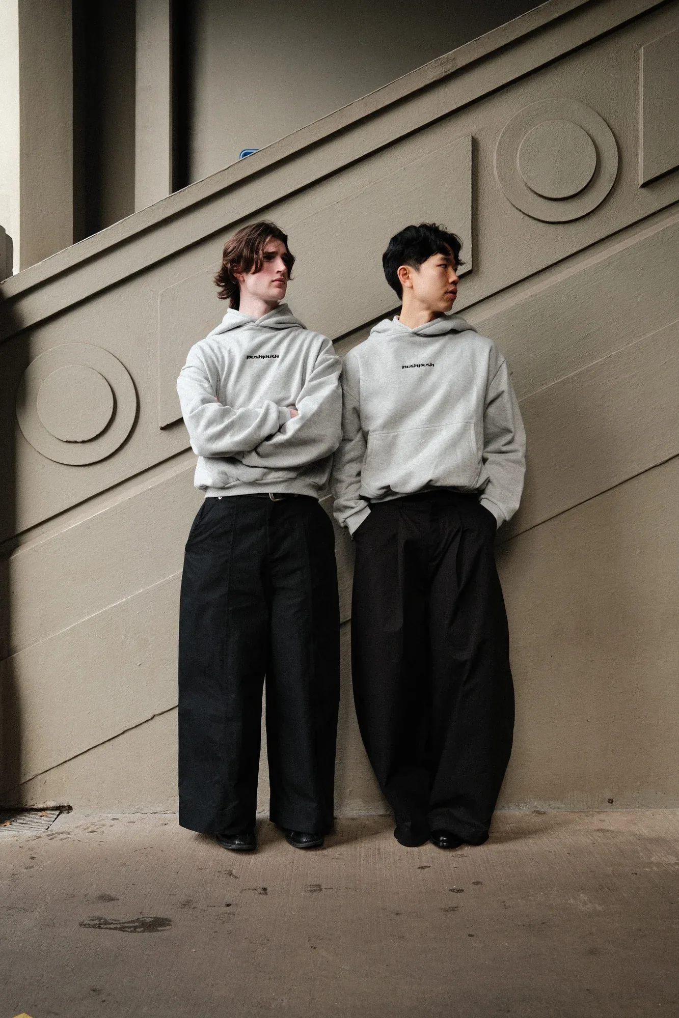 Two models in matching grey hoodies and wide black trousers lean against a carved stone wall
