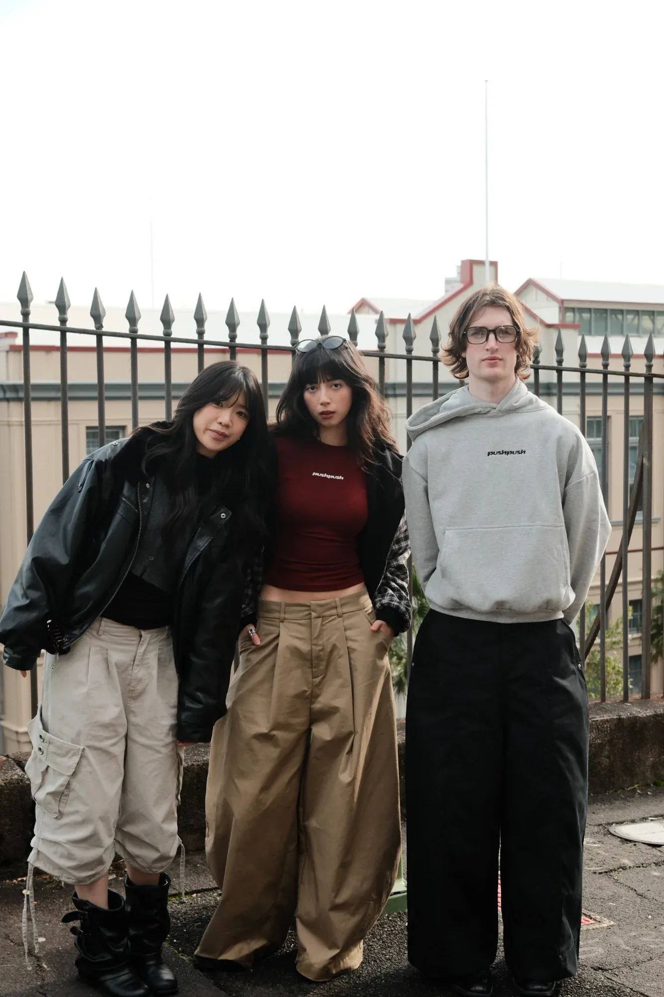 Three models at an iron fence: a leather jacket and cargo pants, a red top and tan wide-leg trousers, a grey hoodie