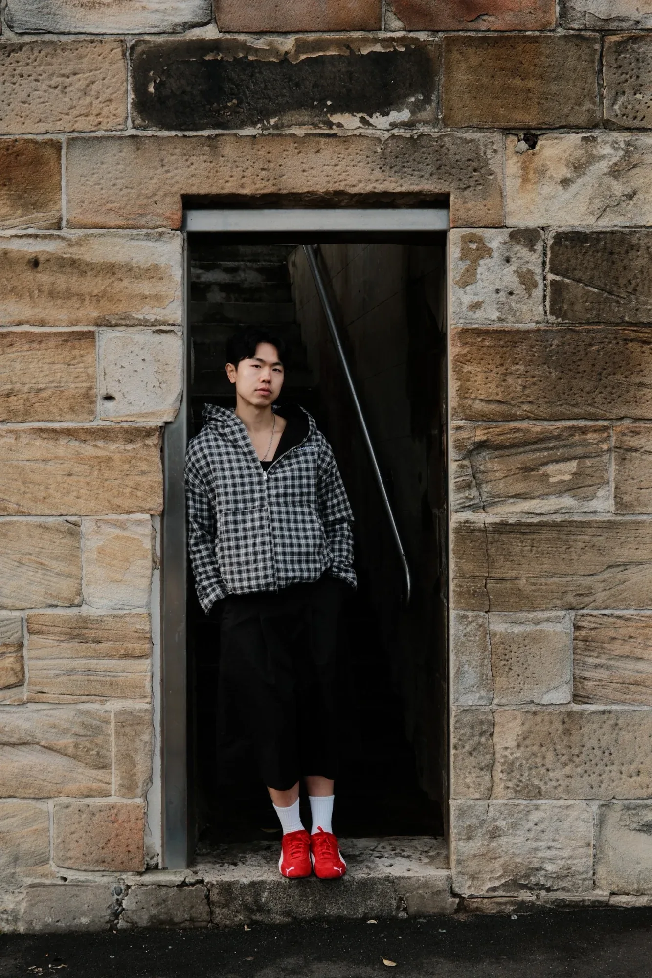 A model in a gingham overshirt and red sneakers stands in a sandstone doorway