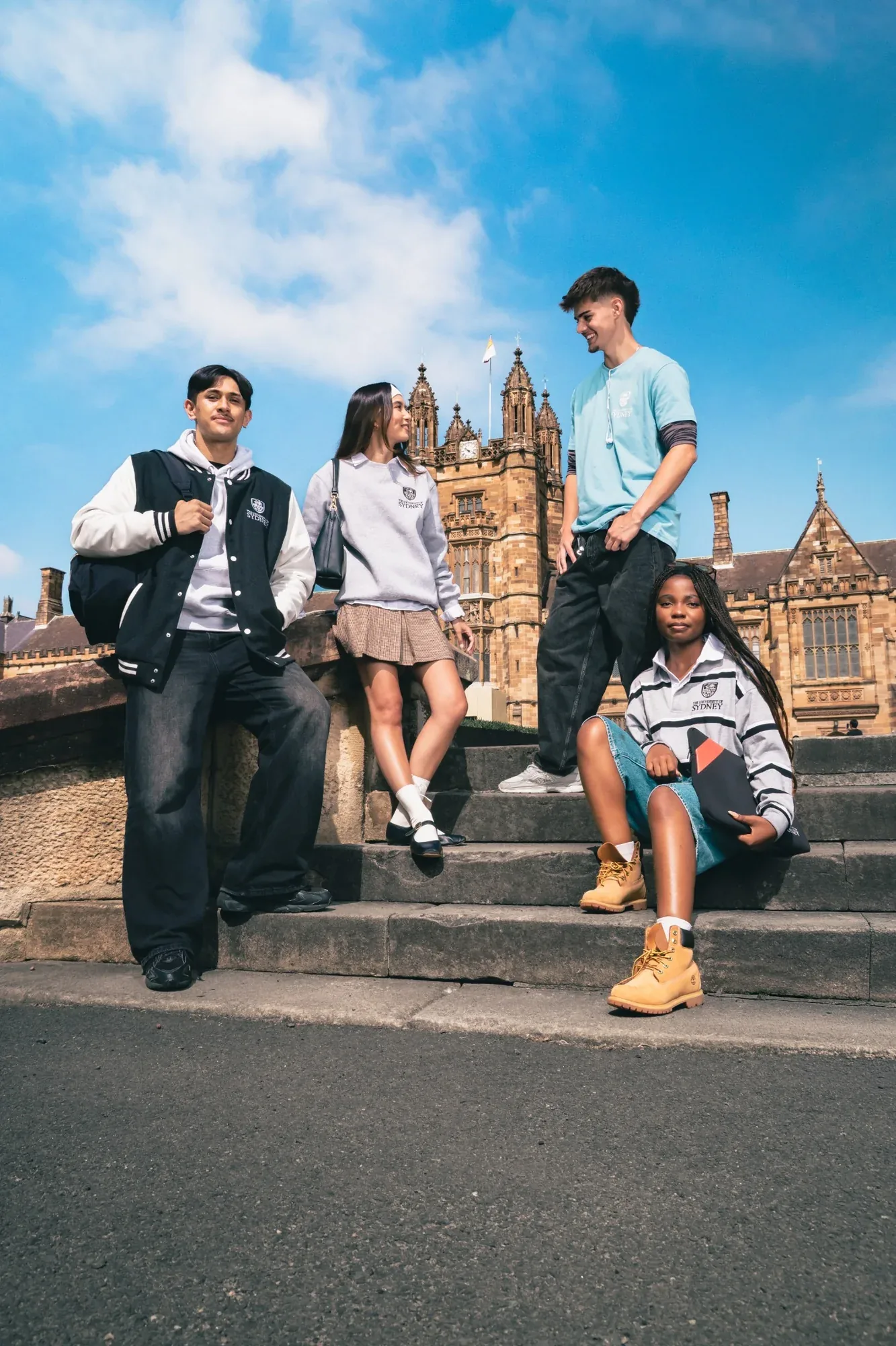 Four students in USU merchandise on the sandstone steps of the University of Sydney Quadrangle under a blue sky