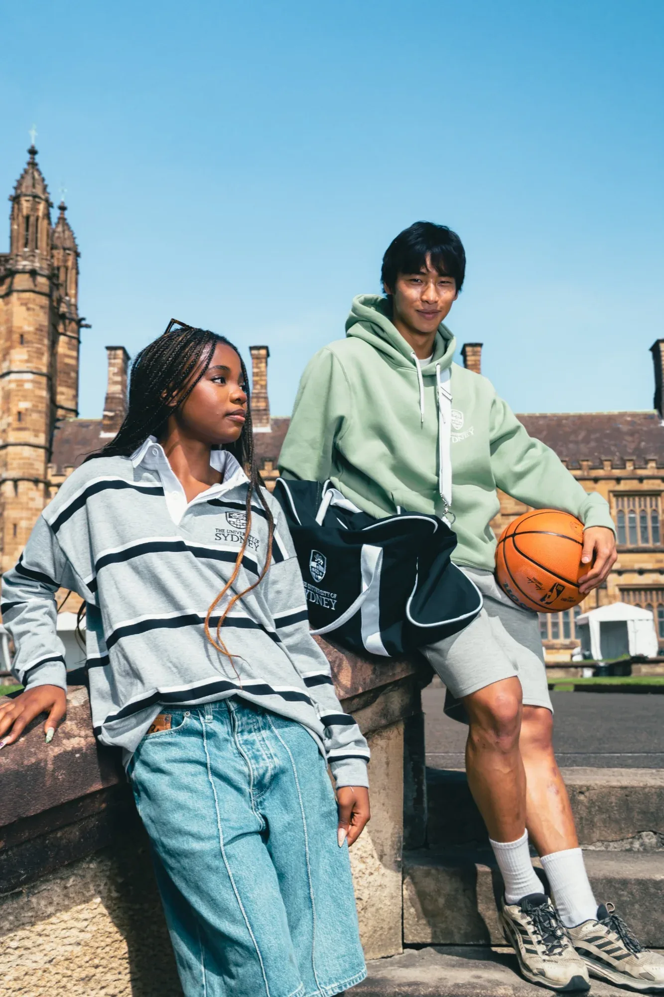 Two students in a USU hoodie and a striped rugby top lean on a sandstone wall, one holding a basketball