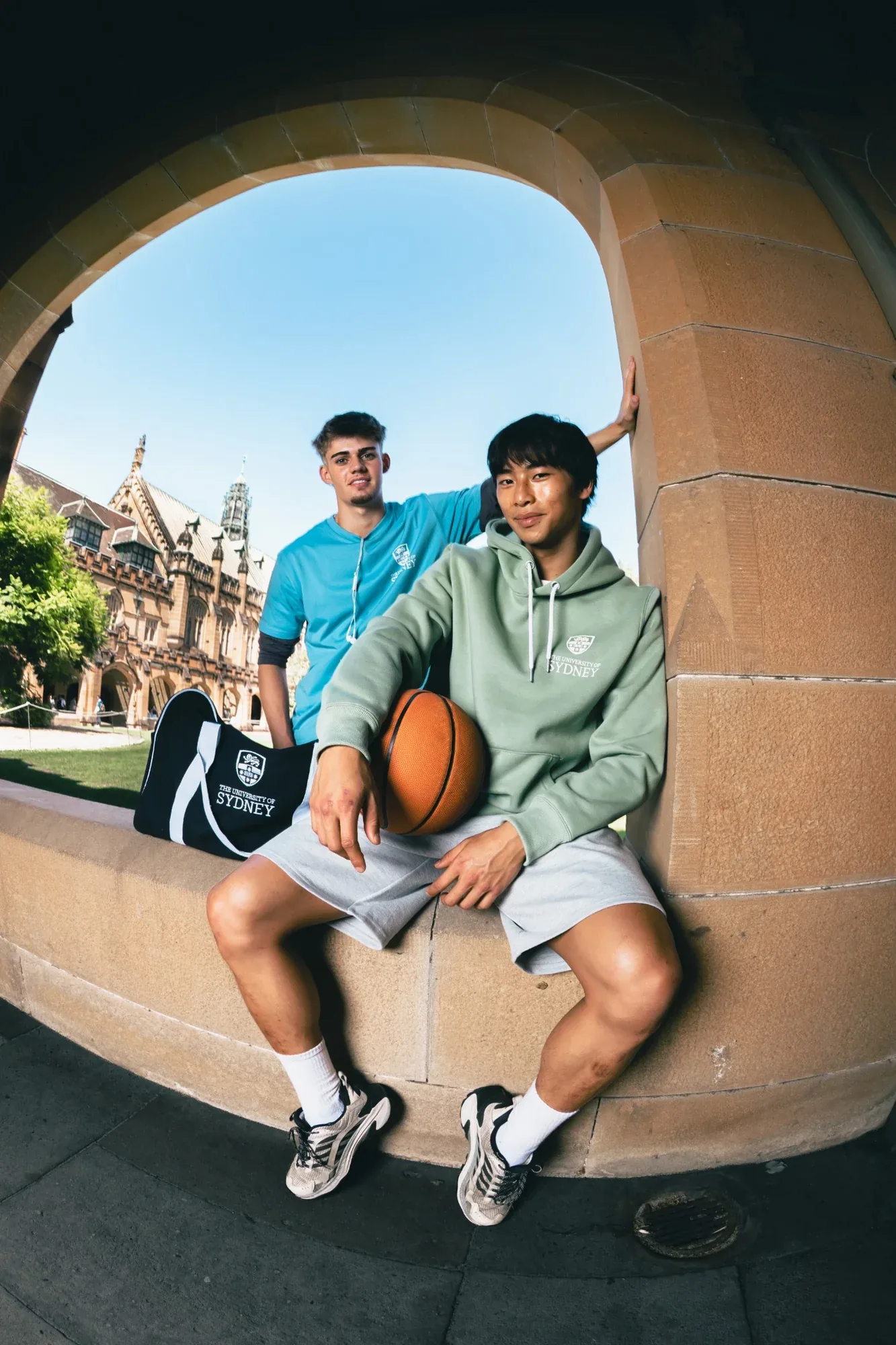 Two students in USU merchandise sit framed in a sandstone archway, one with a basketball