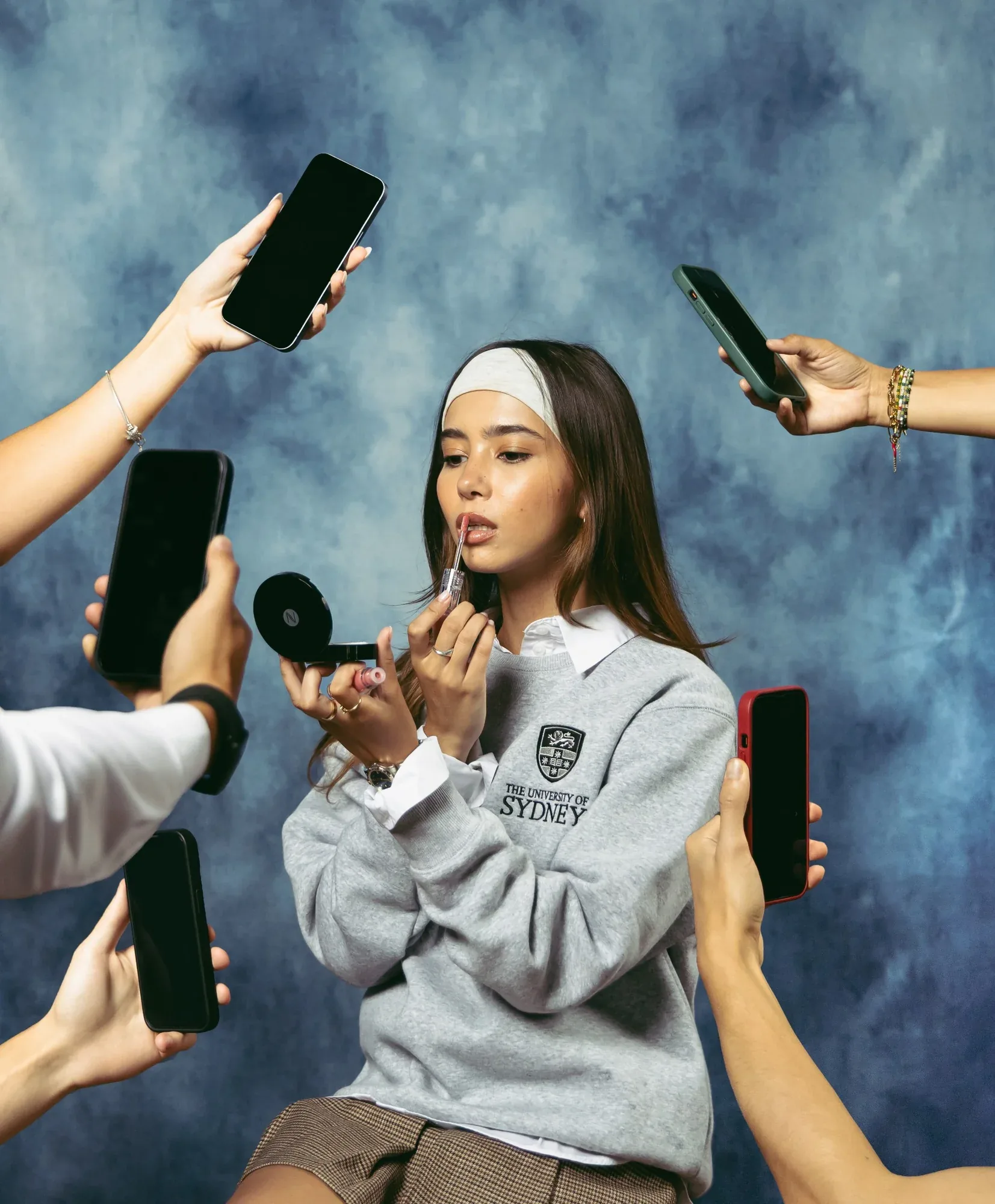 A student in a grey University of Sydney sweater surrounded by hands holding phones, against a painted blue backdrop