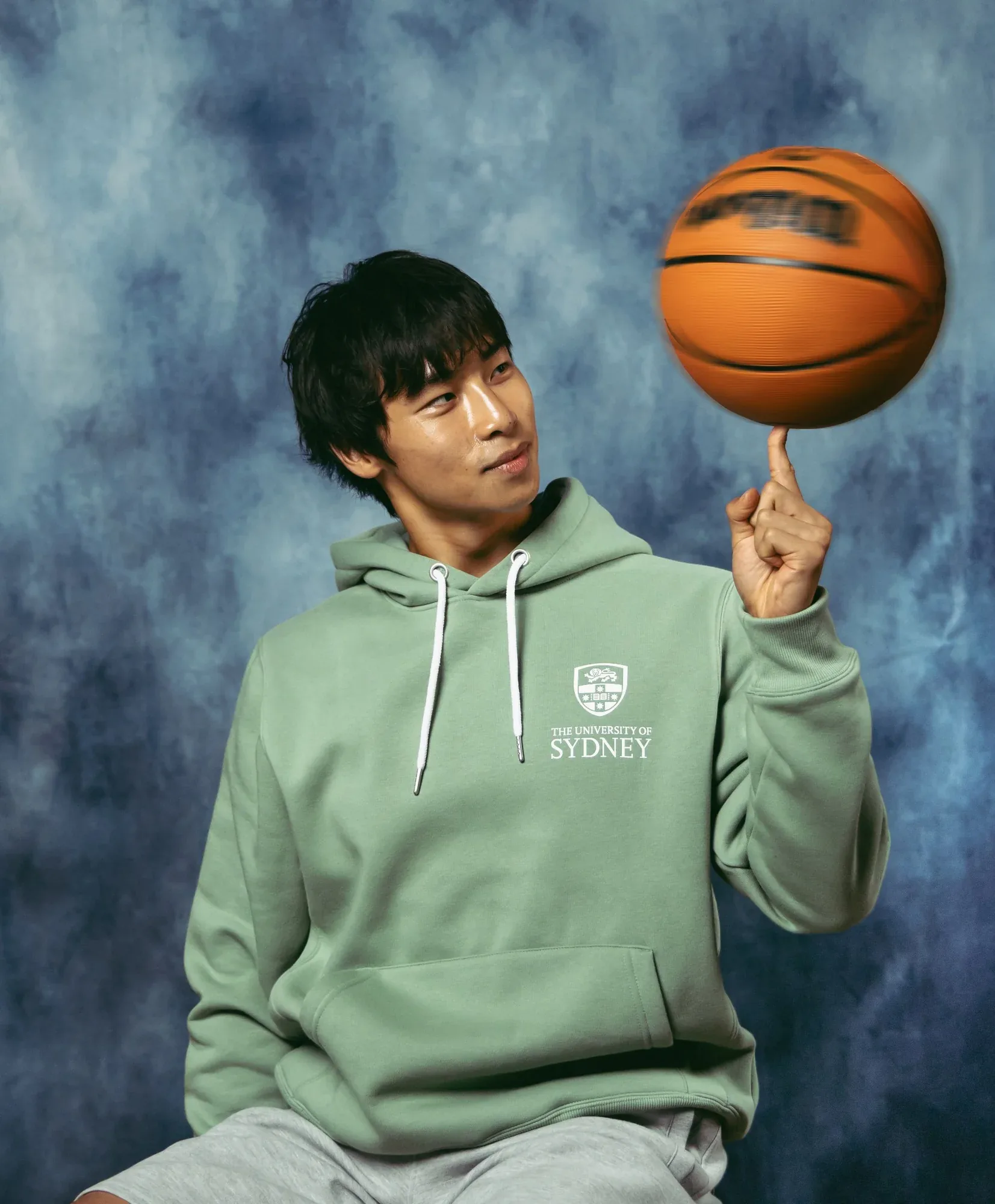 A student in a sage University of Sydney hoodie spins a basketball on one finger against a painted blue backdrop