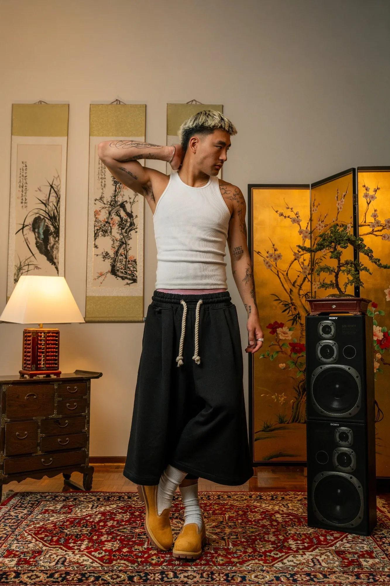A model in a white singlet and wide black shorts poses beside Chinese ink scrolls, a gold screen and a tall speaker