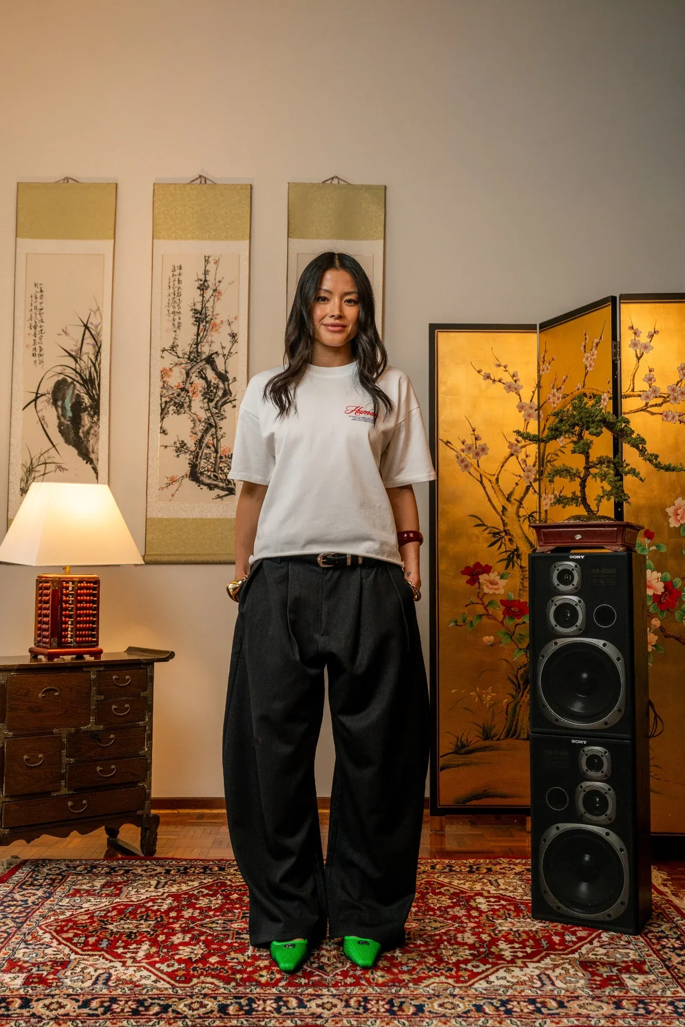 A model in a white tee and wide black trousers stands on a red rug between ink scrolls and a tall speaker