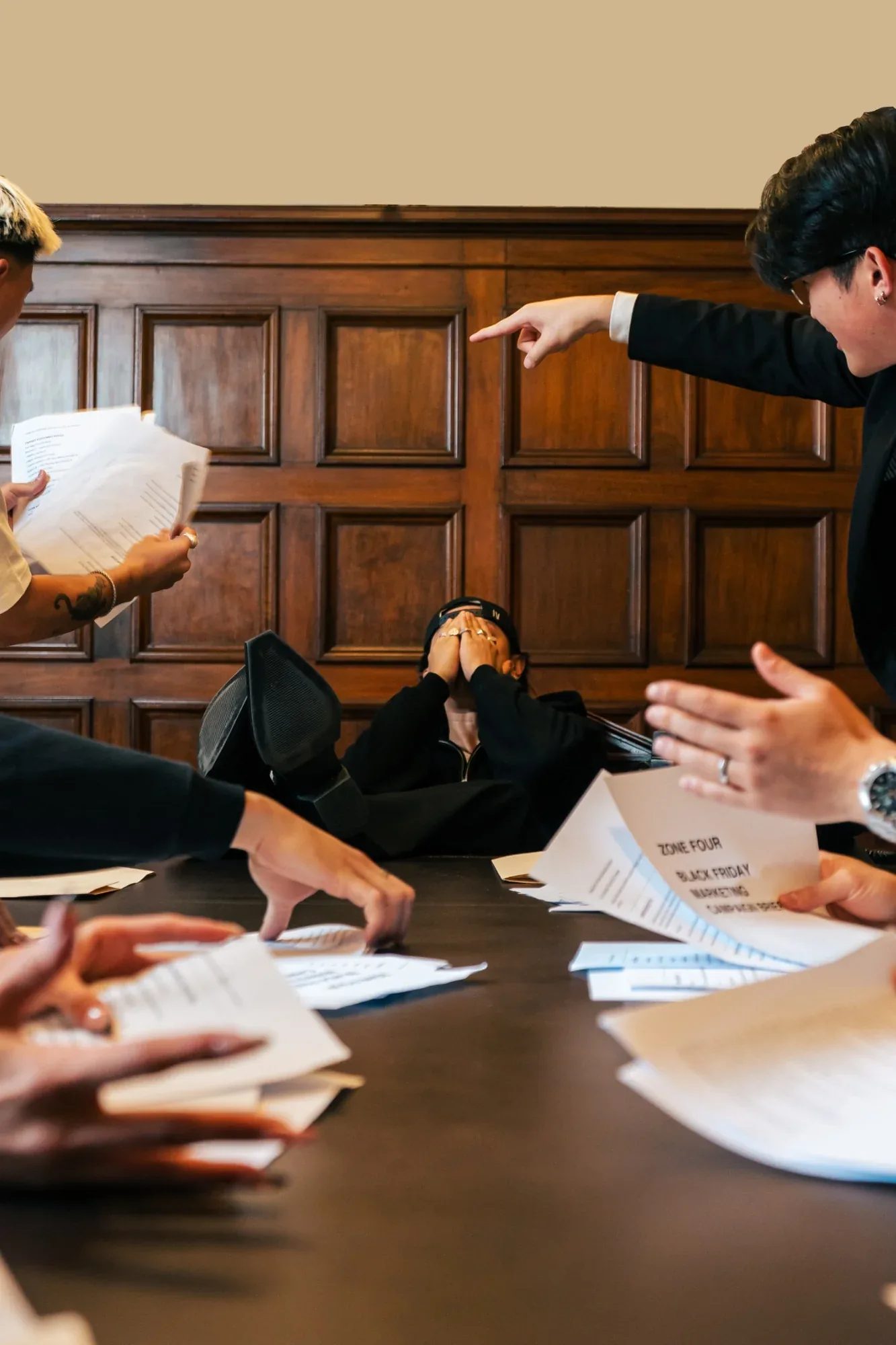 Low angle across a boardroom table as people in suits point and throw papers