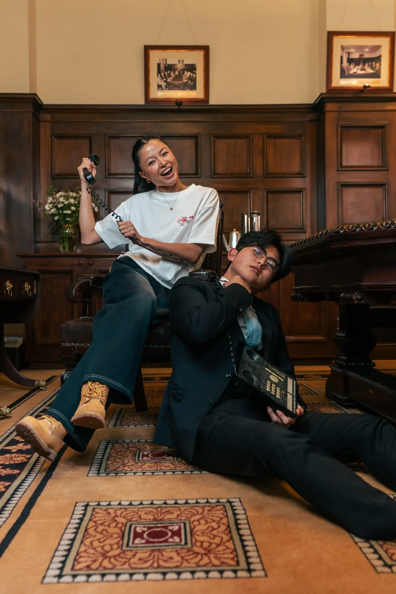One model lounges in a chair while another, in a black suit, slumps on the floor holding a desk phone in a wood-panelled room