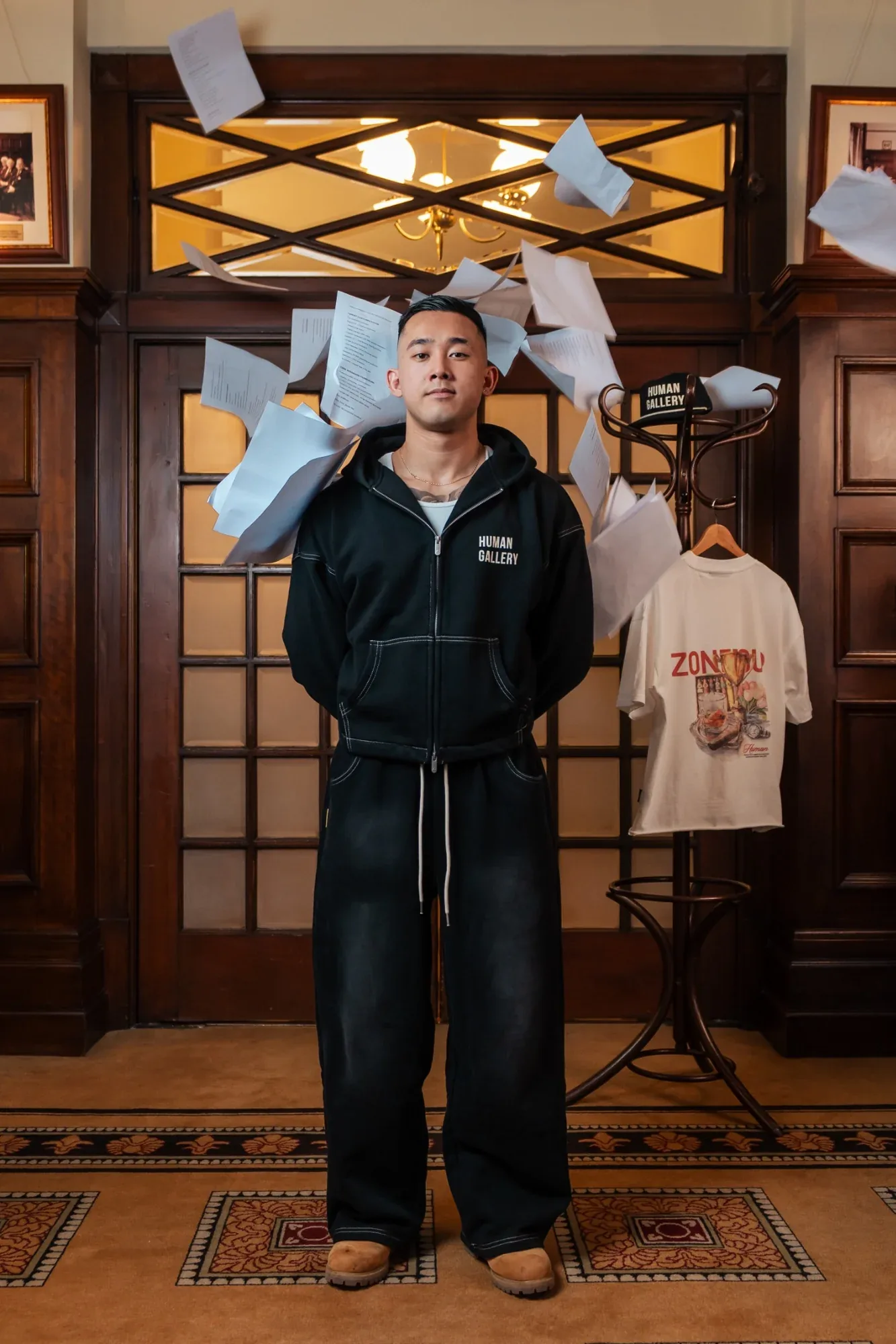 A model in a black Zone Four zip hoodie stands in a wood-panelled doorway as papers fly through the air