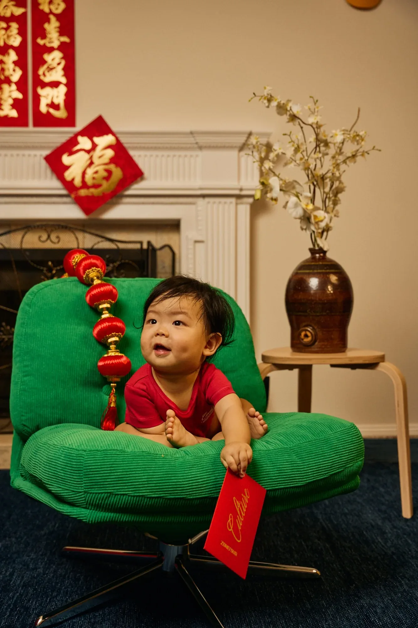 A baby in red holds a red envelope on a green armchair, a fu sign and couplets behind