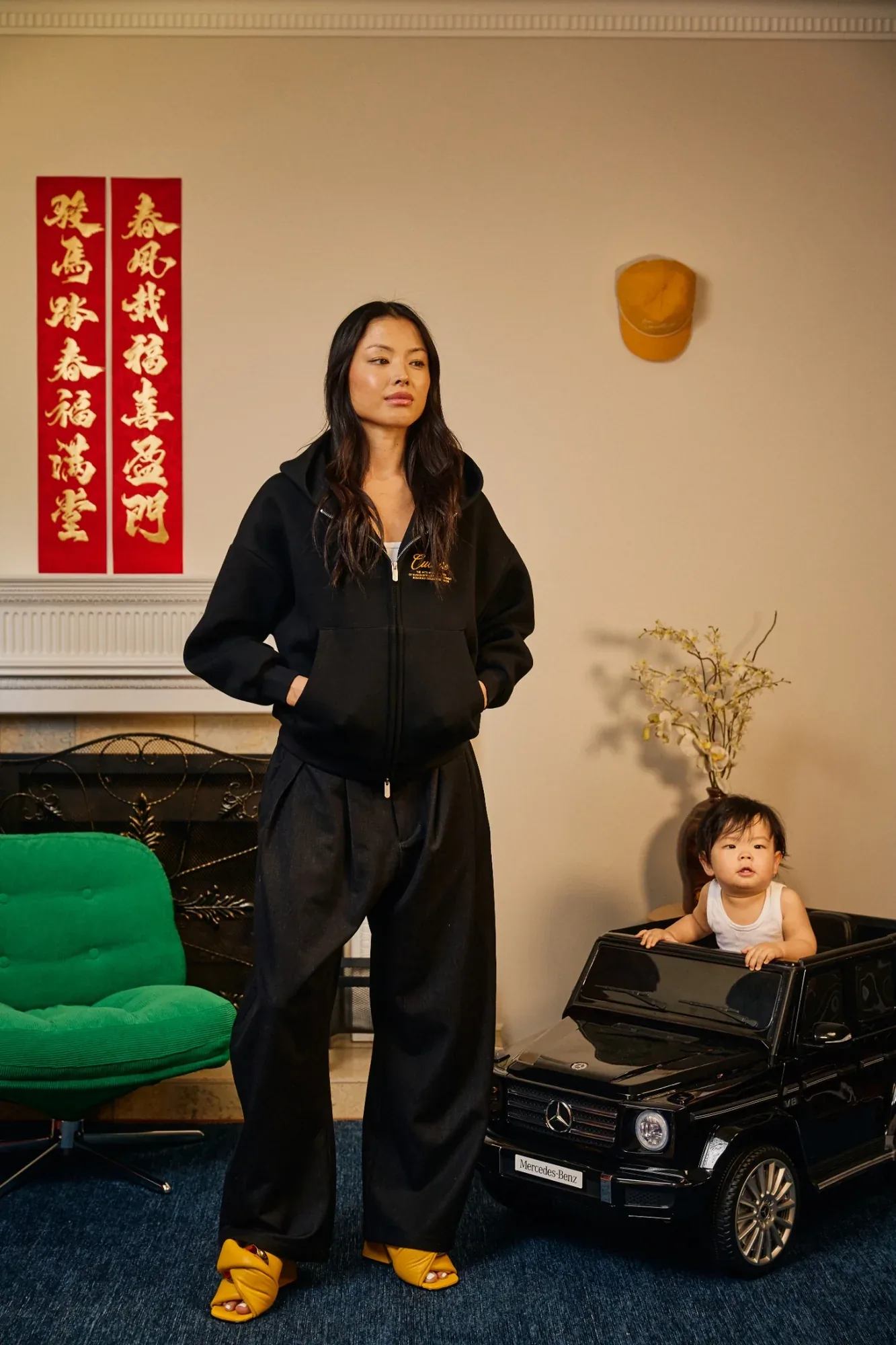 A model in a black Zone Four zip hoodie stands beside a toddler in a toy car, red couplets on the wall