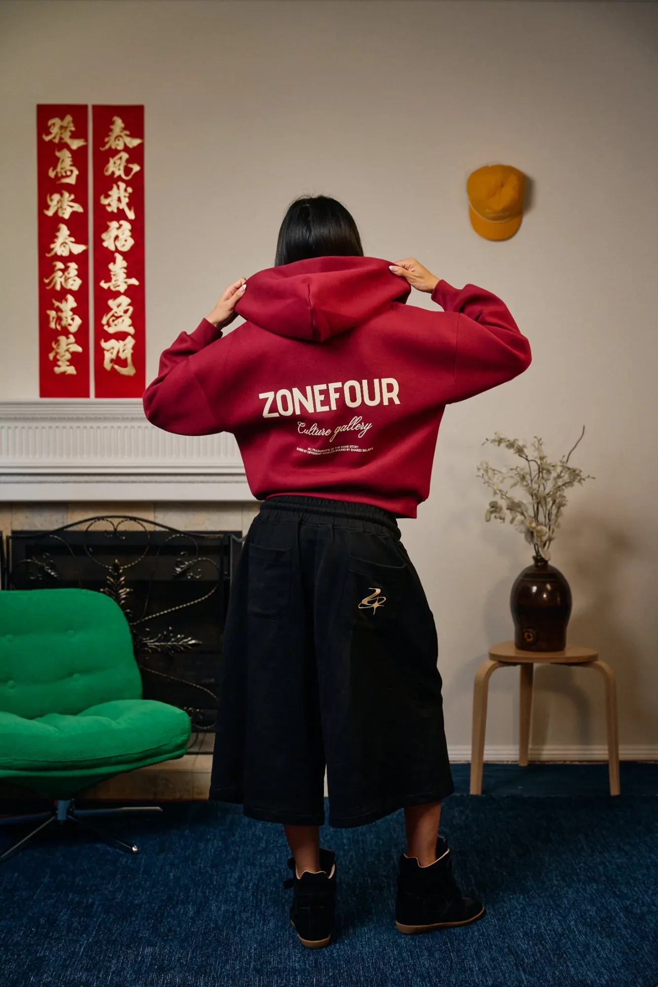 A model seen from behind in a red Zone Four hoodie, hands raised to the hood, red New Year couplets on the wall