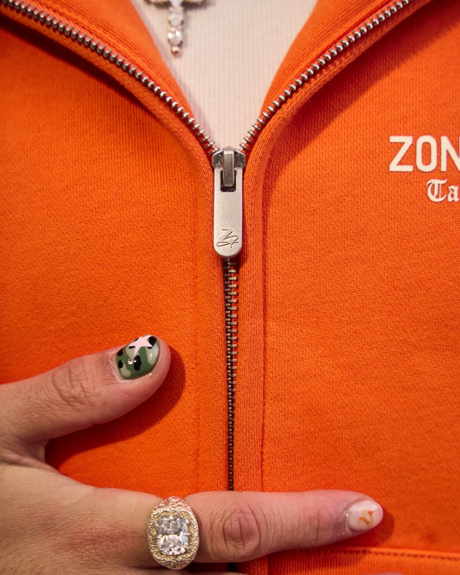 Detail of an orange Zone Four zip hoodie: the branded zip pull, a camo-painted fingernail and a silver ring