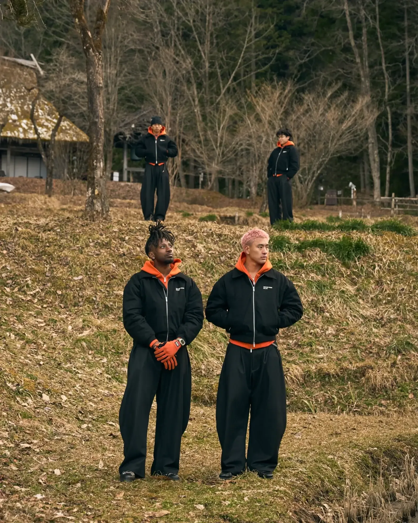 Four models in matching black Zone Four jackets over orange hoodies stand on a grassy slope near a thatched farmhouse