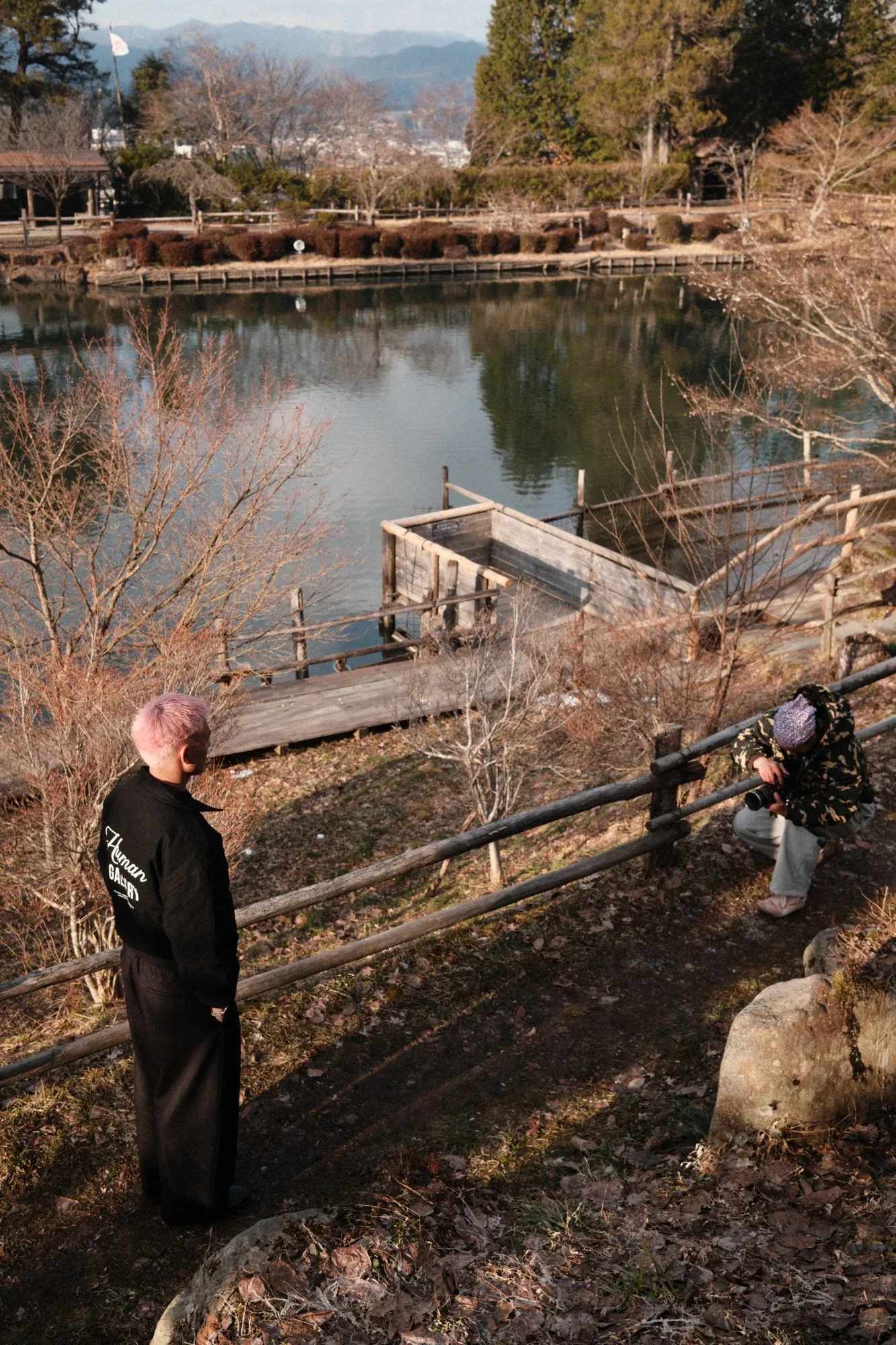 A model with pink hair in a black Zone Four jacket looks out over a lake while a photographer crouches to shoot
