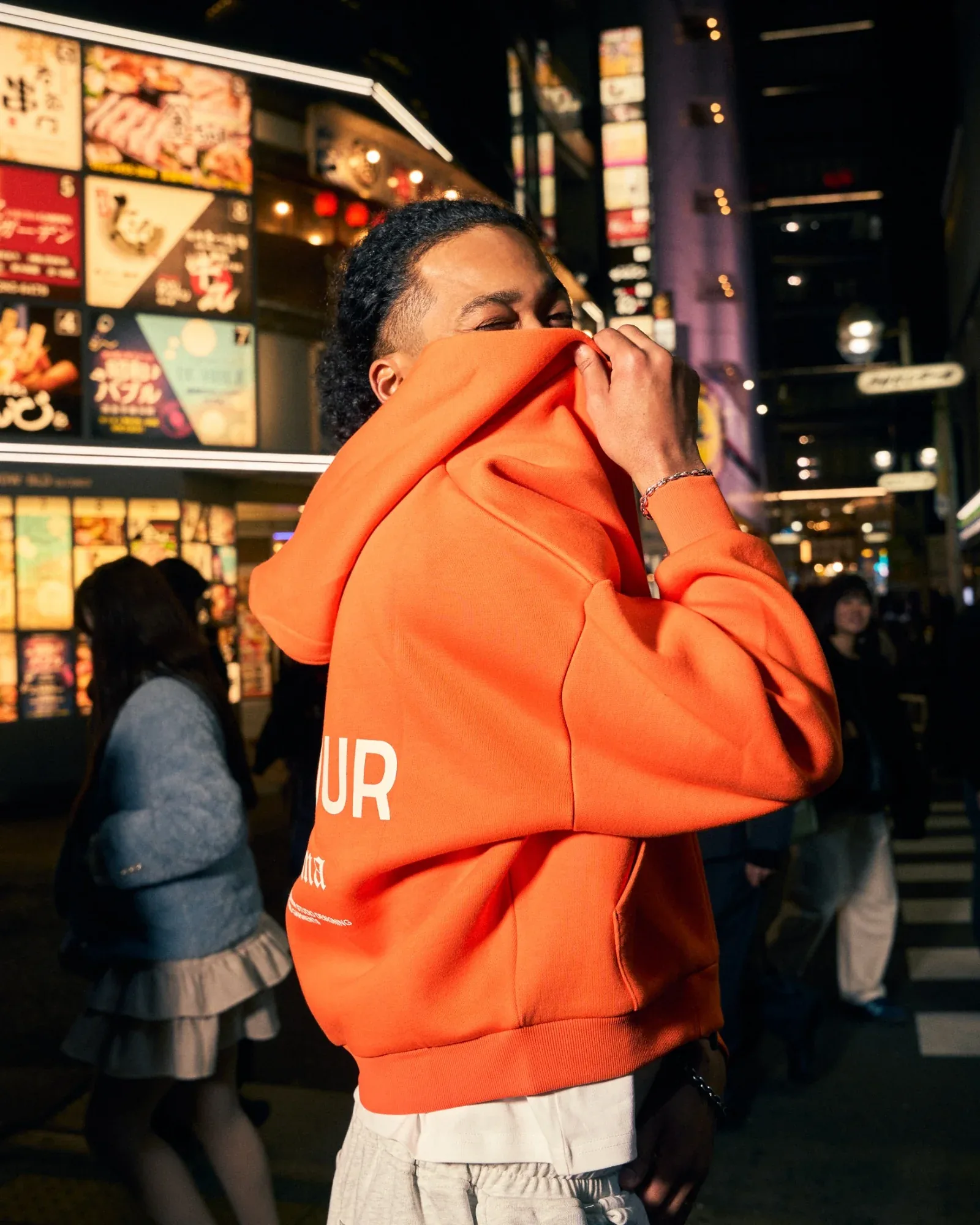A model pulls an orange Zone Four hoodie up over their face on a neon-lit street at night