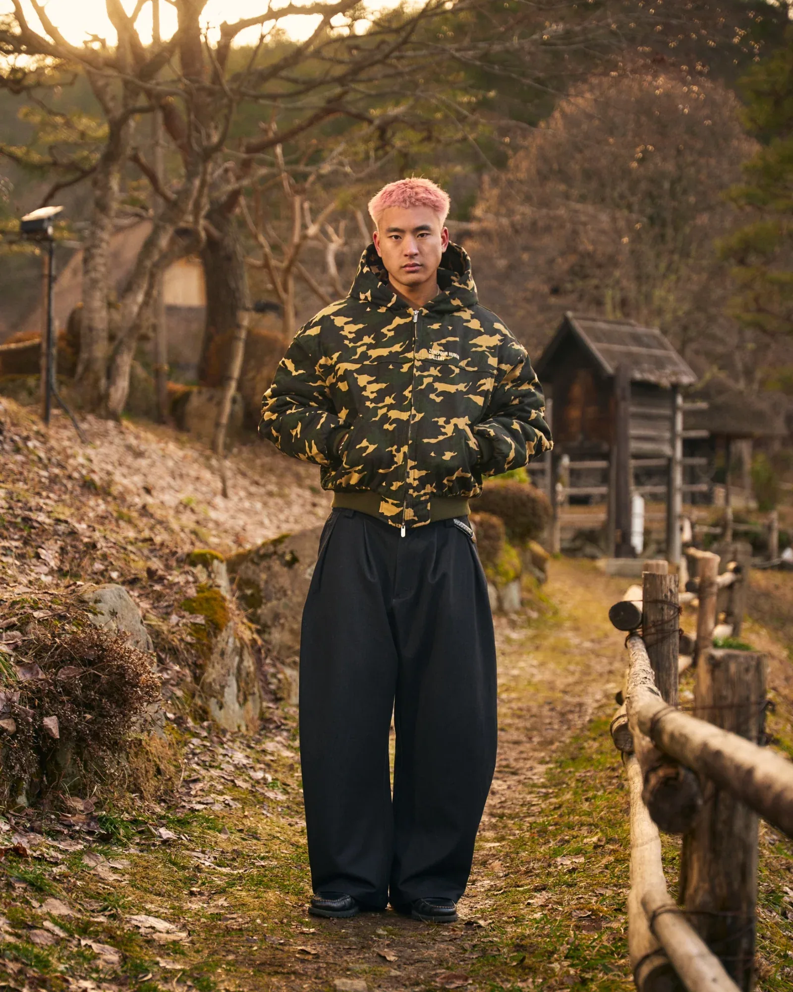 A model with pink hair in the camo hooded bomber and wide black trousers on a leafy path beside a wooden fence