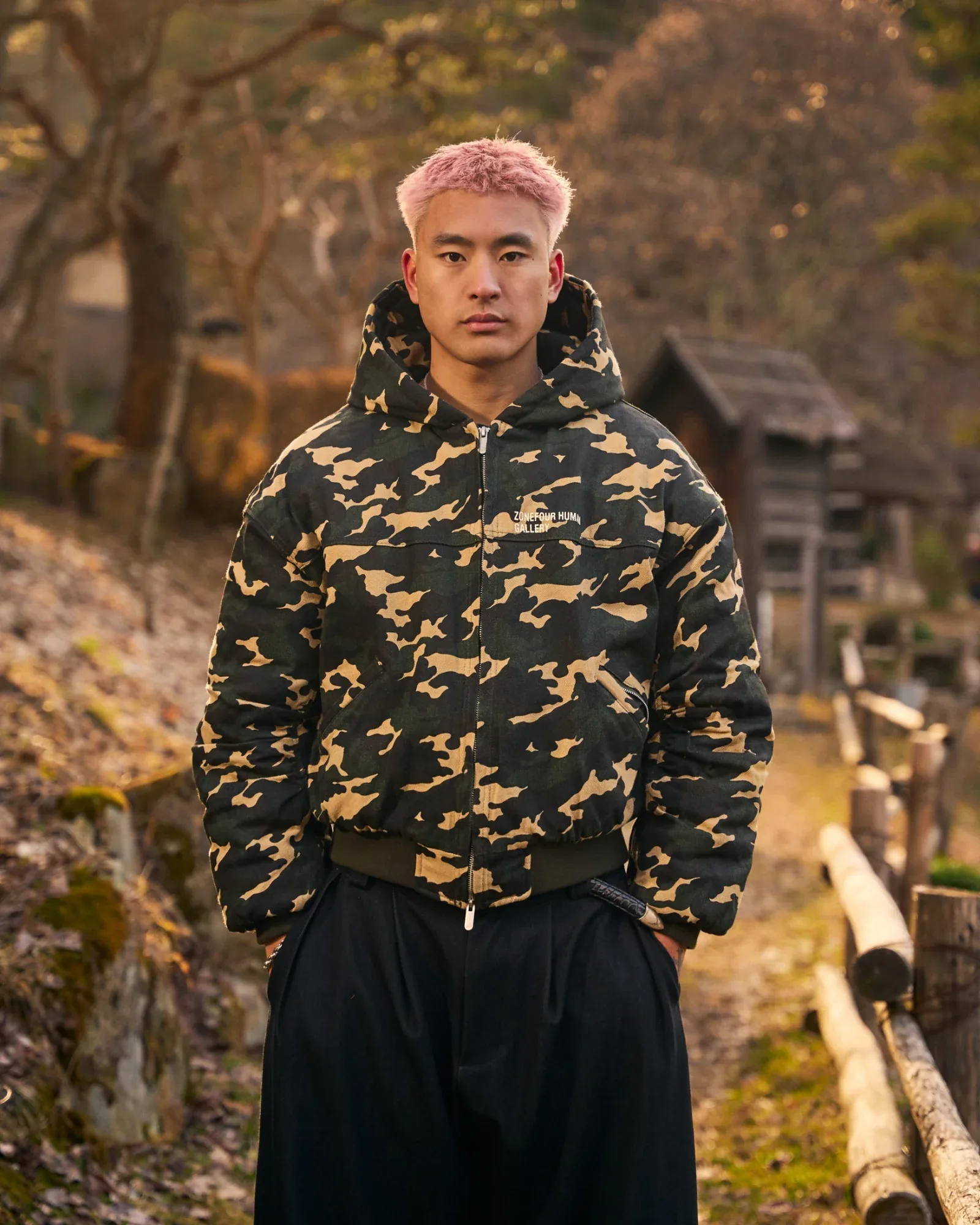 Close portrait of the model in the camo hooded bomber, a thatched hut soft in the background