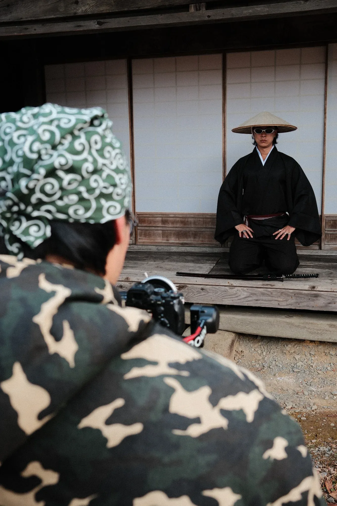 Over the shoulder of a photographer in a camo jacket, framing a figure in black and a straw hat seated on a temple veranda