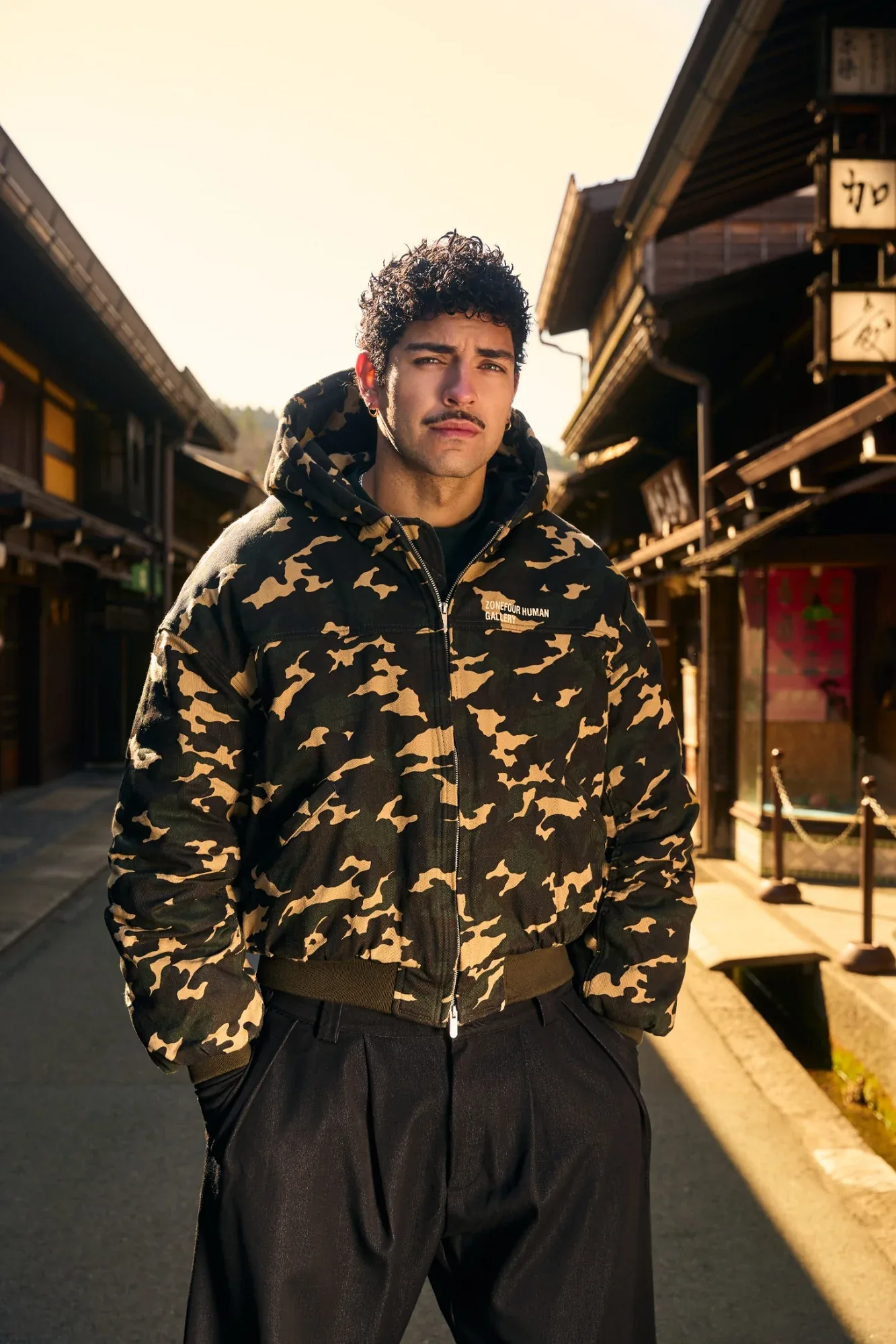A model in a Zone Four Human Gallery camo hooded bomber stands in a street of traditional timber houses, lit by low sun