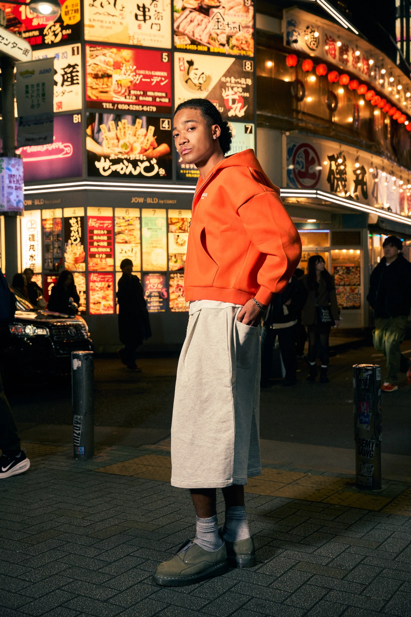 A model in an orange Zone Four hoodie and wide grey shorts stands on a Tokyo street at night, neon restaurant signs behind